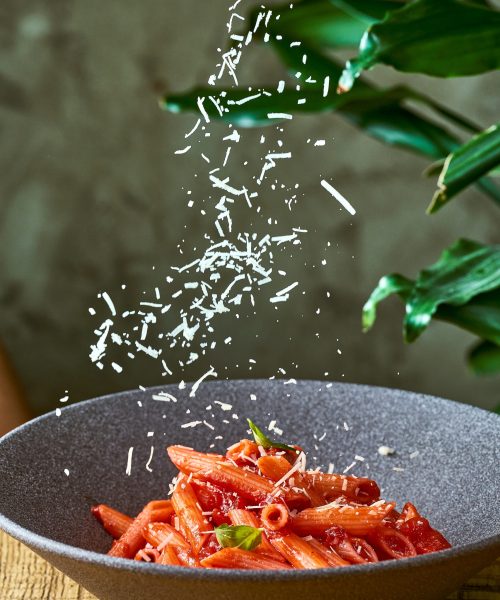 Penne pasta with tomato sauce and basil in a gray plate. Parmesan cheese is falling.